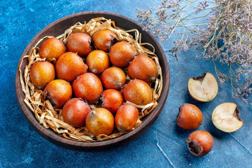 front view of whole and chopped mespilus germanica inside and outside a brown bowl on blue